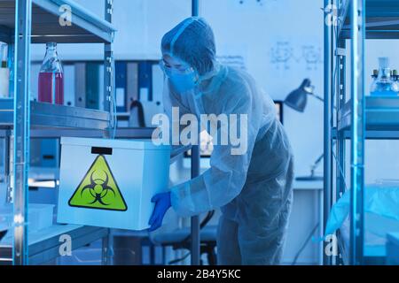 Horizontaler Schuss von nicht erkennbaren medizinischen Wissenschaftlerinnen, die im Laborraum eine Schachtel mit biologischen Gefahren auf das Regal legen Stockfoto
