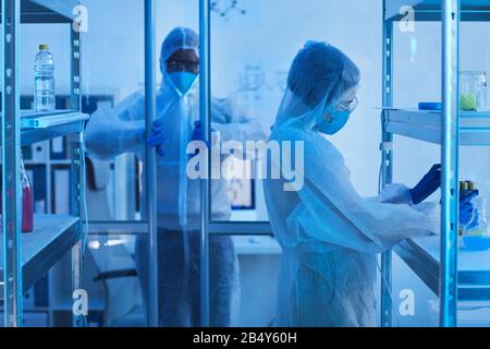 Nicht erkennbare männliche Wissenschaftlerin in schützender Arbeitskleidung, die in den Laborraum eindringt, seine Kollegin nimmt die Reagenzgläser vom Regal Stockfoto