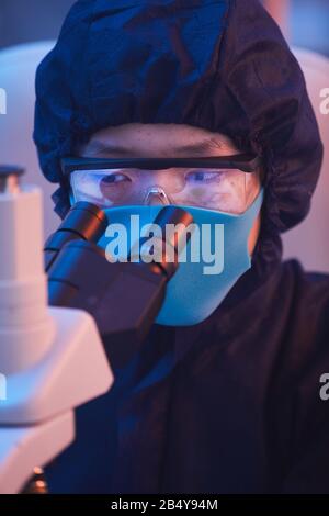 Vertikaler Nahaufnahme von nicht erkennbaren jungen Menschen, die Laborschutzanzug, Brille und Maske tragen, mit dem Mikroskop, um Virus zu untersuchen Stockfoto