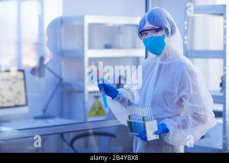 Nicht erkennbare weibliche Pharmakologin trägt einen Einweg-Schutzschlauch mit gleichmäßigem Halteschlauch mit blauer Flüssigkeit, die auf die Kamera blickt, horizontales mittleres Sho Stockfoto