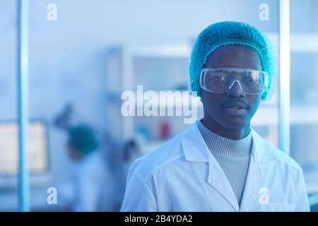Horizontaler Kopf und Schultern Porträt des gutaussehenden jungen Mannes mit weißem Labormantel, medizinischer Kappe und Schutzbrille mit Blick auf die Kamera Stockfoto