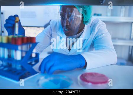 Horizontaler mittlerer Nahaufnahme des jungen männlichen Laborwissenschaftlers Black, der das Reagenzglas wählt und herausnimmt Stockfoto