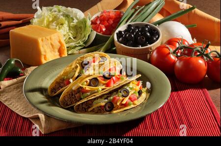 Drei Tacos auf einem Teller, umgeben von Zutaten. Die Farbpalette ist traditionell mexikanisch. Stockfoto