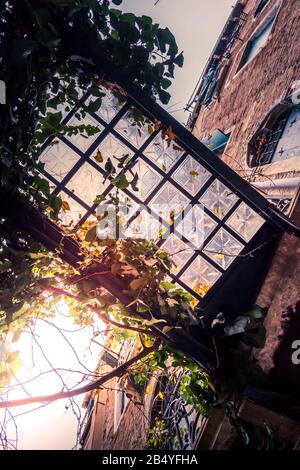 Abstrakt aufgenommen von unten, einer verzierten Glasfliese und einer eisernen Fußgängerbrücke in einer Hintergasse von Venedig, Italien, während das Sonnenlicht die Wolken durchbricht Stockfoto