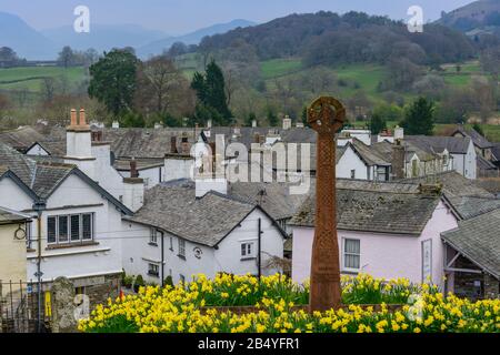 Ein von Narzissen umgebenen Denkmal auf dem Gelände der St Michaels und Der All Angels Church mit dem Dorf Hawkshead, Cumbria, im Hintergrund. Stockfoto