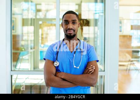 Porträt eines ernsthaften jungen männlichen Arztes oder einer Krankenschwester mit blauen Schrubben Uniform und Stethoskop, mit im Krankenhaus gekreuzten Armen Stockfoto
