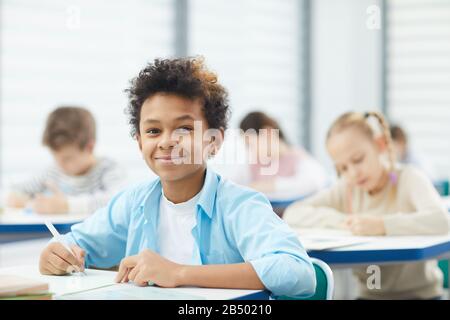 Horizontales Brustbild des gutaussehenden gemischten Jungen mit weißem T-Shirt und blauem Hemd, das am Schultisch sitzt und die Kamera betrachtet Stockfoto
