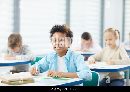 Horizontales mittleres Nahporträt des fröhlichen gemischten Jungen mit knabrigen Haaren am Schulschalter, der lächelnd auf die Kamera blickt und Platz für Kopien bietet Stockfoto