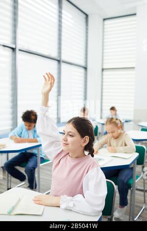 Vertikales Porträt des aktiven, eleganten zwölfjährigen Mädchens mit dunklem Haar, das am Schultisch sitzt und die Hand in der Klasse hebt, Platz für Kopien Stockfoto