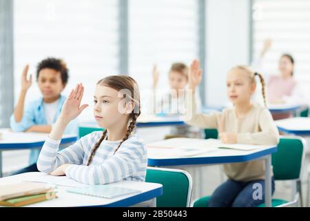 Aktive junge Jungen und Mädchen, die an Schultischen sitzen und ihre Hände in der Klasse heben, horizontaler Schuss, Kopierraum Stockfoto