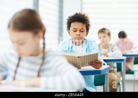 Horizontales mittleres Porträt des zwölfjährigen Jungen, der am Schreibtisch im modernen Lesebuch im Klassenzimmer sitzt, Kopierer Stockfoto