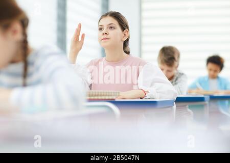 Horizontales Porträt des aktiven, intelligenten zwölfjährigen Mädchens, das am Schulschalter sitzt und die Antwort geben möchte, indem sie ihre Hand in der Klasse, im Kopierraum, hochhebt Stockfoto