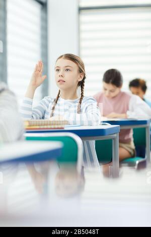 Vertikales Porträt der jungen Schulmädchensitzerin und des Schreibtischs, die die Hand heben und ihrem Lehrer während der Unterrichtszeit Fragen stellen, Platz kopieren Stockfoto