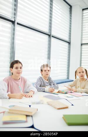 Vertikaler Schuss von jungen und zwei Mädchen, die im modernen Klassenzimmer sitzen und den Lehrern zuhören, Kopierraum Stockfoto