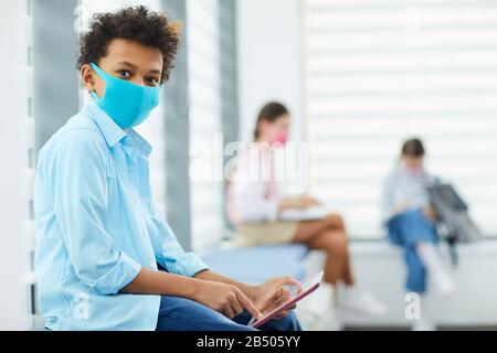 Horizontales Porträt eines nicht erkennbaren schwarzen Jungen, der eine Maske trägt, die in einer Queque sitzt, um in den medizinischen Räumen der Schule zu sitzen, in denen das Gerät die Kamera und den Kopierraum betrachtet Stockfoto