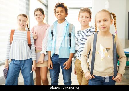Porträt von zwei Jungen und drei Mädchen, die auf dem Schulflur zusammen stehen und auf der Kamera posieren, horizontaler mittlerer langer Schuss Stockfoto