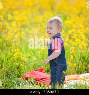 Kleiner Baby-Junge, der im Frühjahr auf der Wiese mit Löwenzahn Spaß hat. Wunderschönes lächelndes Kind auf einem grünen Feld mit gelben Blumen in der Natur. Stockfoto