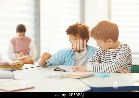 Horizontales Porträt von zwei männlichen Schülern der Mittelschule, die am Schreibtisch im regnerischen Klassenzimmer sitzen und gemeinsam Aufgaben erledigen, Platz kopieren Stockfoto