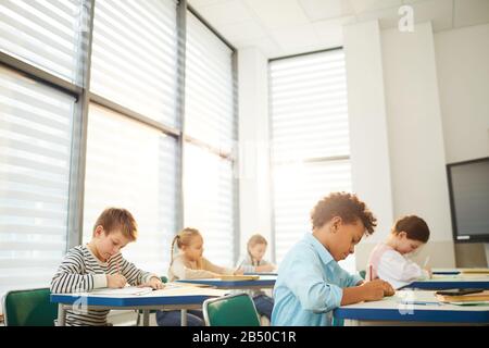 Horizontale Aufnahme junger Mittelschüler, die an Schreibtischen im modernen Klassenzimmer sitzen und an Unterricht arbeiten, Kopierraum Stockfoto