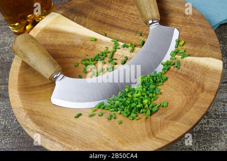 Zubereitung von Kräutern zum Essen: Isoliertes braunes Holzschneidebrett mit dem Zerkleinerungsmesser des Küchenchefs, gehackte grüne Schnittlauch Stockfoto