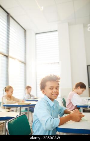 Vertikales Porträt eines fröhlichen Mixed-Race-Jungen mit knabrigen Haaren am Schulschalter, der lächelnd auf die Kamera blickt und Platz für Kopien bietet Stockfoto