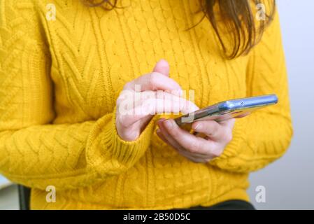 Smartphone in den Händen einer Frau in einem gelben warmen Pullover, ein Konzept zum Thema Dating in sozialen Netzwerken, Einkaufen im Internet Stockfoto