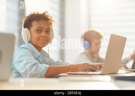 Mittleres Porträt eines afroamerikanischen Jungen, der Kopfhörer mit Laptop während des Studiums in der Schule trägt, Kopierraum Stockfoto