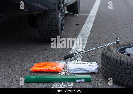 Erste Hilfe, orangefarbene Weste und grünes Straßenschild auf einer Asphaltstraße vor dem Hintergrund eines zerbrochenen Autos. Notfall-Werkzeugsatz für die erste Hilfe nach dem Auto Stockfoto