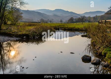 Im April spiegelte sich die untergehende Sonne in River Rothay vor dem Dorf Grasmere, Lake District National Park, Cumbria, England, Großbritannien, wider. Stockfoto