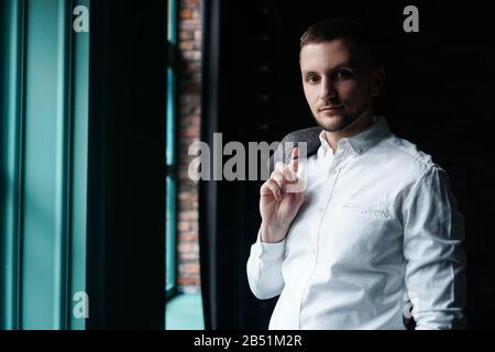 Blick in das Profil eines jungen Geschäftsmannes, gekleidet mit einem weißen Hemd, das in der Nähe des Fensters an einer dunklen Wand steht und die Kamera betrachtet Stockfoto