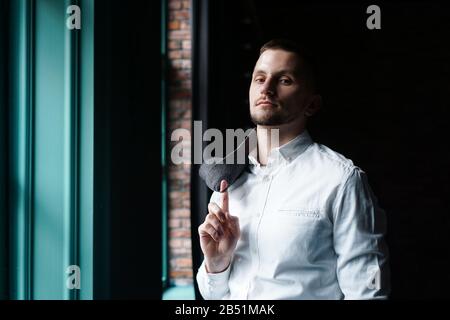Blick in das Profil eines jungen Geschäftsmannes, gekleidet mit einem weißen Hemd, das in der Nähe des Fensters an einer dunklen Wand steht und die Kamera betrachtet Stockfoto