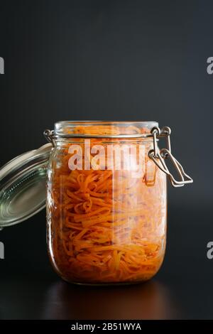 500 Gramm würziger koreanischer Karottensalat mit Öl, Knoblauch und schwarzem Pfeffer im Glasbecher. Nachhaltige Verpackung, schwarzer Hintergrund, hohe Auflösung Stockfoto