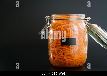 500 Gramm würziger koreanischer Karottensalat mit Öl, Knoblauch und schwarzem Pfeffer im Glasbecher. Nachhaltige Verpackung, schwarzer Hintergrund, hohe Auflösung Stockfoto