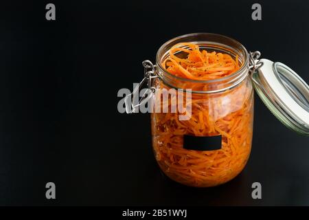 500 Gramm würziger koreanischer Karottensalat mit Öl, Knoblauch und schwarzem Pfeffer im Glasbecher. Nachhaltige Verpackung, schwarzer Hintergrund, hohe Auflösung Stockfoto