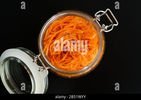 500 Gramm würziger koreanischer Karottensalat mit Öl, Knoblauch und schwarzem Pfeffer im Glasbecher. Nachhaltige Verpackung, schwarzer Hintergrund, hohe Auflösung Stockfoto