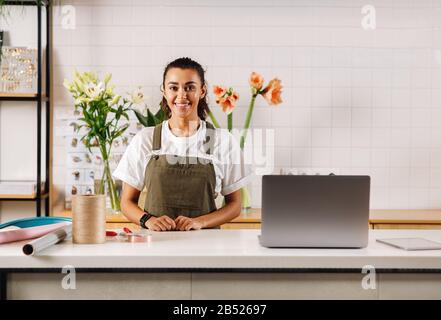 Lächelnde Frau in der Schürze, die an der Theke steht und die Kamera betrachtet. Schöner Blumenhändler im Blumenladen. Stockfoto