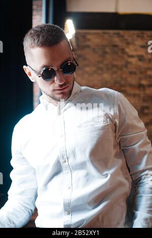 Gutaussehender junger Mann mit einer runden schwarzen Vintage-Brille, die in der Nähe der roten Ziegelwand nach vorne blickt Stockfoto