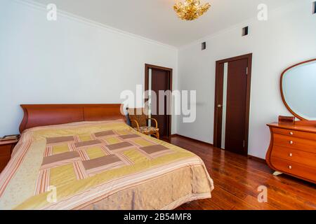 Ein großes Schlafzimmer mit Mahagoni, Korbstühlen und einem eleganten Schminktisch mit einem halbkreisförmigen Spiegel. Das Bett hat eine beige Decke in Quadraten. Stockfoto