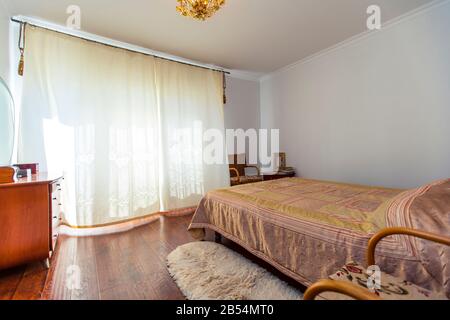 Ein großes Schlafzimmer mit Mahagoni, Korbstühlen und einem eleganten Schminktisch mit einem halbkreisförmigen Spiegel. Das Bett hat eine beige Decke in Quadraten. Stockfoto