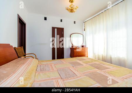 Ein großes Schlafzimmer mit Mahagoni, Korbstühlen und einem eleganten Schminktisch mit einem halbkreisförmigen Spiegel. Das Bett hat eine beige Decke in Quadraten. Stockfoto