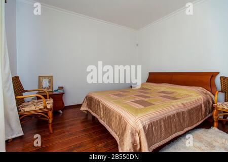 Ein großes Schlafzimmer mit Mahagoni, Korbstühlen und einem eleganten Schminktisch mit einem halbkreisförmigen Spiegel. Das Bett hat eine beige Decke in Quadraten. Stockfoto