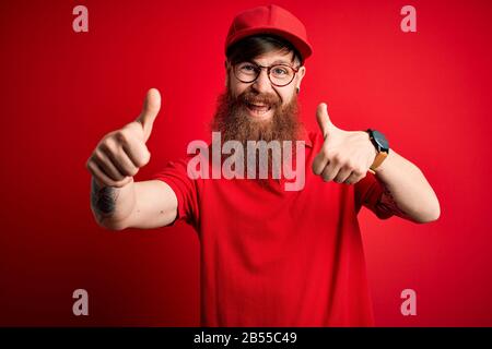 Junger, gutaussehender Liefermann, der eine Brille und eine rote Kappe über dem isolierten Hintergrund trägt und positive Geste mit der Hand abzeichnet, mit den Daumen nach oben lächelnd und hap Stockfoto