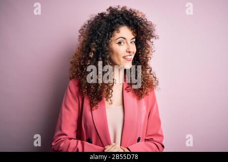 Junge, schöne Geschäftsfrau mit lockigen Haaren und durchstechenden, eleganten Jacke mit Hand und gekreuzten Fingern, die entspannt und fröhlich lächeln Stockfoto