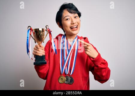 Junge schöne asiatische Mädchen-Gewinnerin, die Trophäe mit Medaillen über weißem Hintergrund trägt, sehr glücklich, mit Hand und Finger zu zeigen Stockfoto