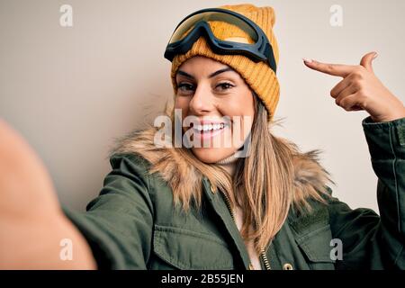 Junge Brunette-Skifahrerin mit Schneekleidung und Skibrille macht selfie mit der Kamera sehr glücklich, mit Hand und Finger zu zeigen Stockfoto