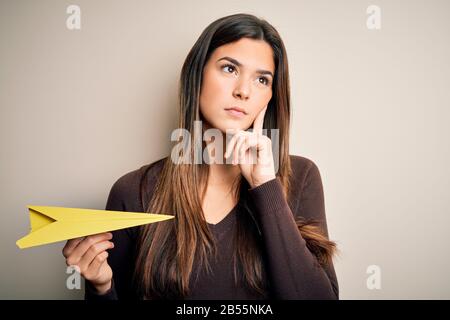 Junges, wunderschönes Mädchen, das Papierflugzeug über einem isolierten weißen Hintergrund hält ernstes Gesicht, das über die Frage denkt, sehr verwirrte Idee Stockfoto