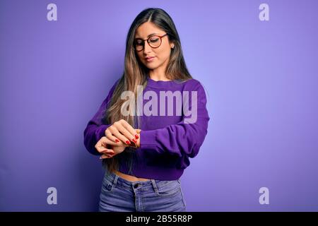 Junge schöne, elegante Frau mit Brille über purpurfarbenem, isoliertem Hintergrund Überprüfen der Zeit auf der Armbanduhr, entspannt und selbstsicher Stockfoto