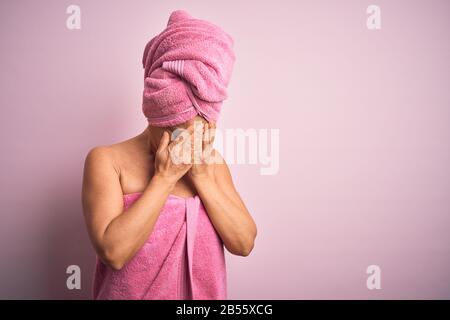 Frau im mittleren Alter, die Badetuch von der Körperpflege über rosafarbenen Hintergrund trägt, mit trauriger Ausdrucksweise, die Gesicht mit den Händen bedeckt, während sie weint. Depression c Stockfoto