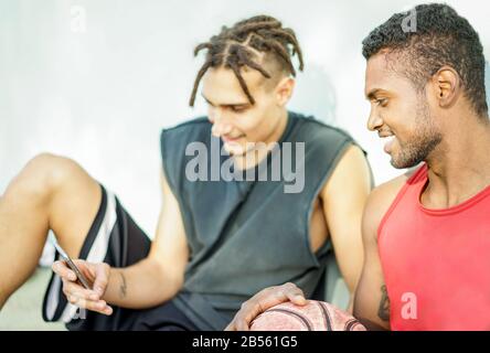 Mehrere ethnische glückliche Freunde, die sich nach dem Basketballtraining entspannen, sehen ein Smartphone aus - Junge Leute, die Spaß an der Universität haben - Mobile Device Suchting Co Stockfoto