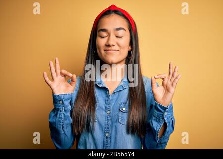 Junge schöne asiatische Frau mit legeren Jeanshemd und diadem über gelbem Hintergrund entspannen Sie sich und lächeln Sie mit geschlossenen Augen, die Meditationsgeste w ausführen Stockfoto